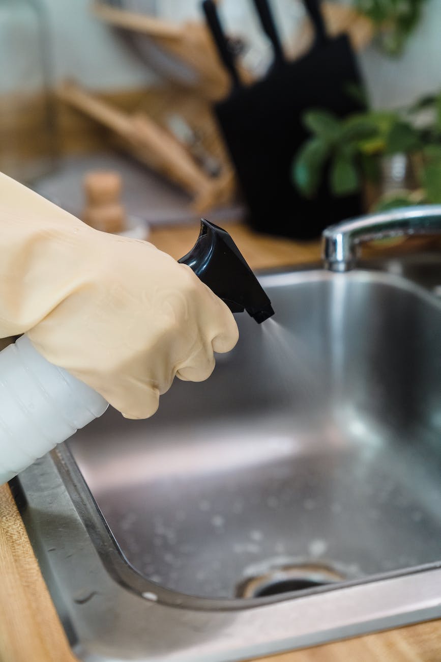 A member of the Steamins team, wearing rubber gloves, is using a spray bottle to apply cleaning products to a stainless steel kitchen sink.