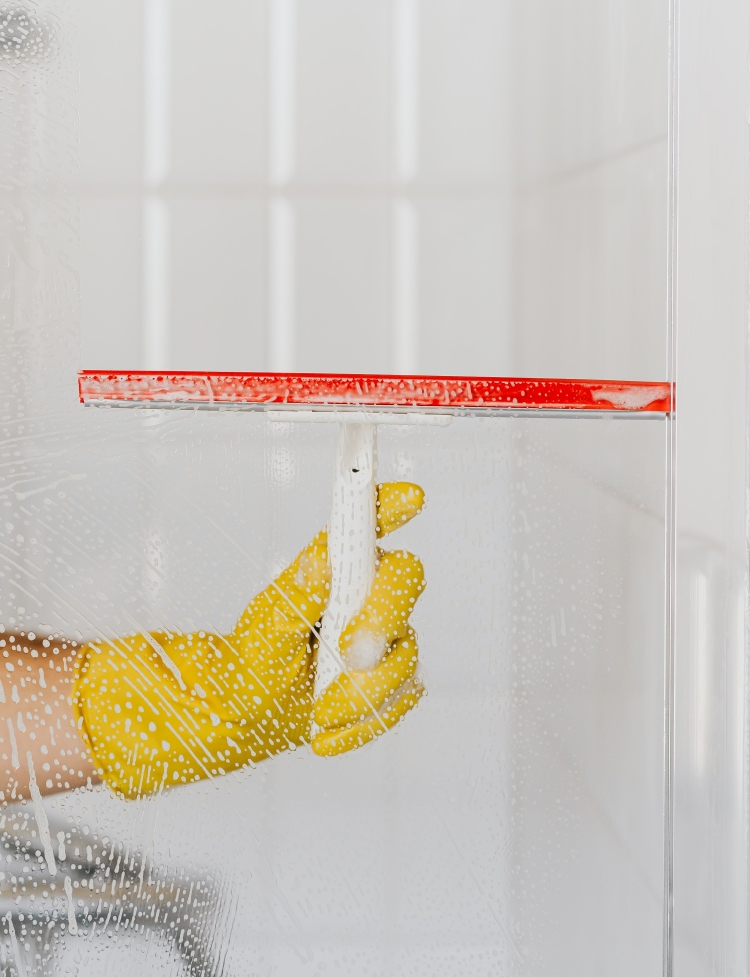 A shower screen is being cleaned by a cleaner in Maiden Bradley, who is wearing gloves and using a squeegee and cleaning solution in a professional manner.